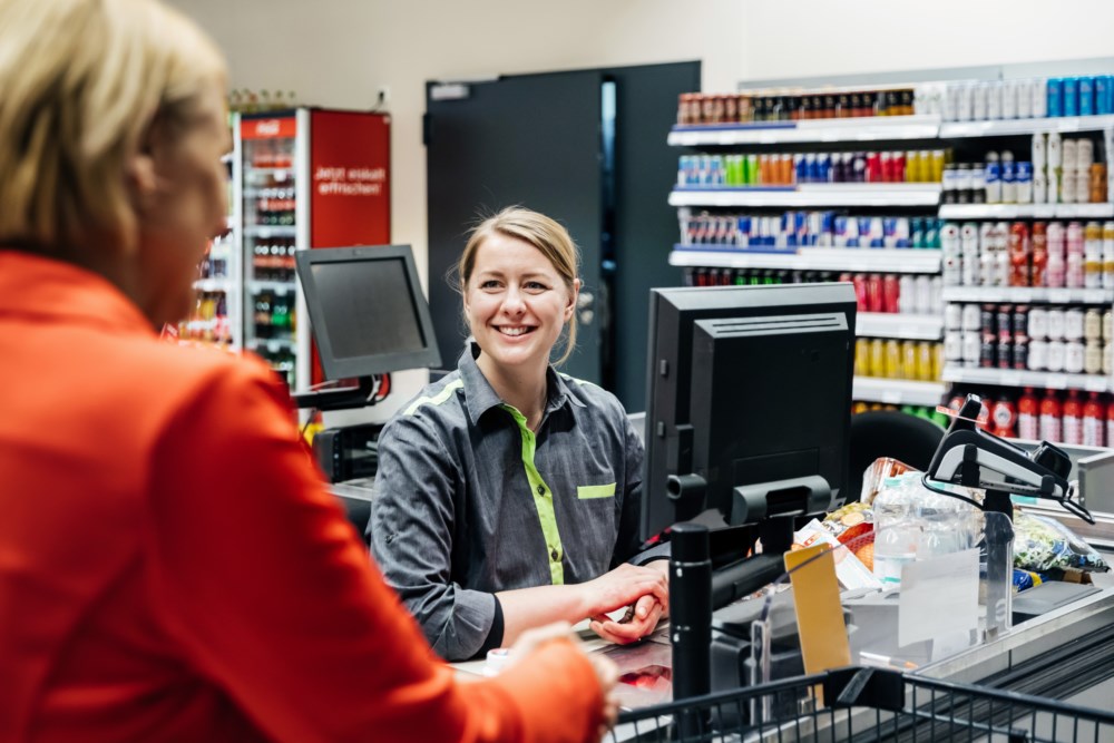 Voksen kvinne ved kassen i dagligvarebutikk, håndterer kontanter ved en bankterminal.